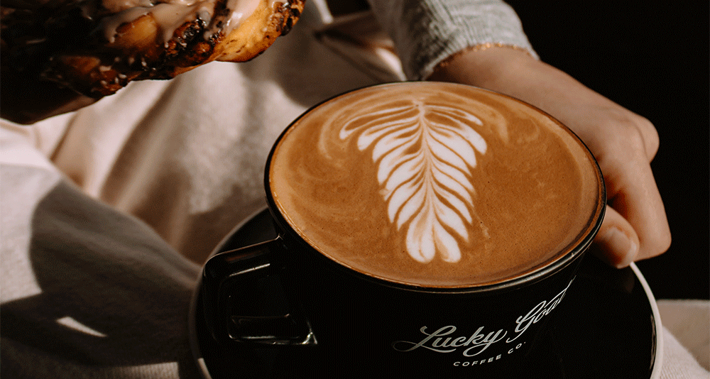 Close up shot of a hot latte in a black mug with saucer, adorned with beautiful latte art. There is a hand holding the saucer and the edge of a pastry in the photo.