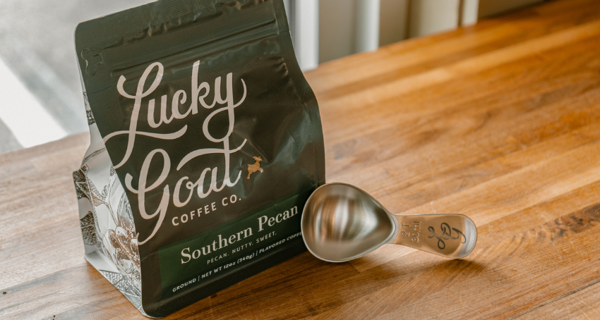 Picture of bag of Southern Pecan Flavored Coffee with a silver LG coffee scoop leaning against the bottom of it on a wooden table in a café.