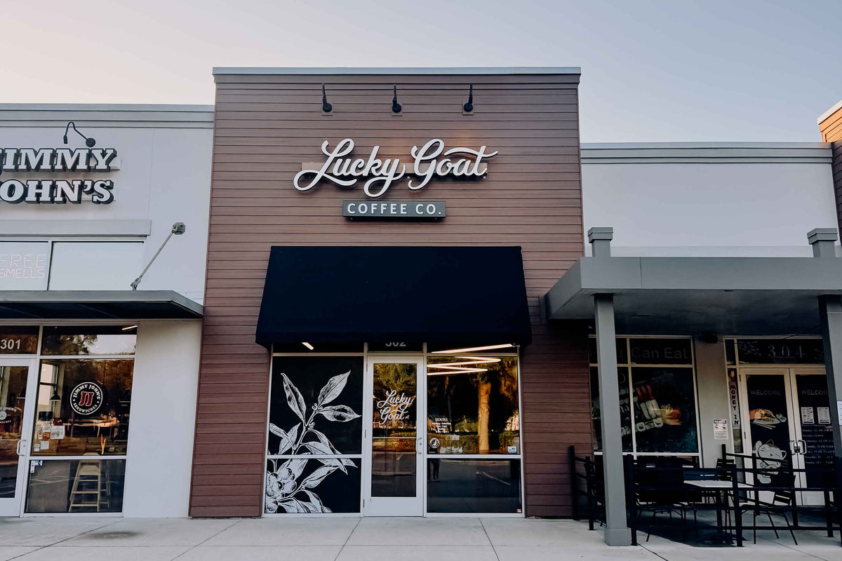 Lucky Goat Coffee Co. storefront with a brown wood-look facade, white script logo, black awning, and glass entrance doors, shown in a strip mall setting.