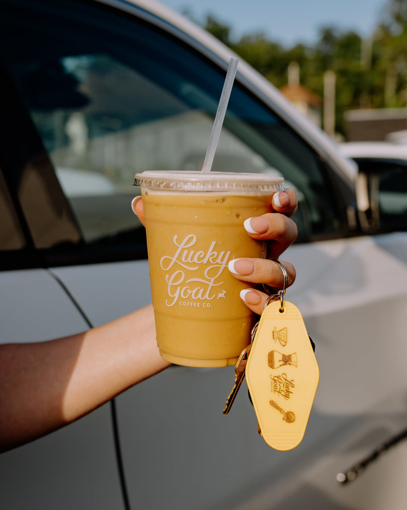 Person holding a cup of coffee with 'Lucky Goat' branding and a yellow keychain, standing next to a car.