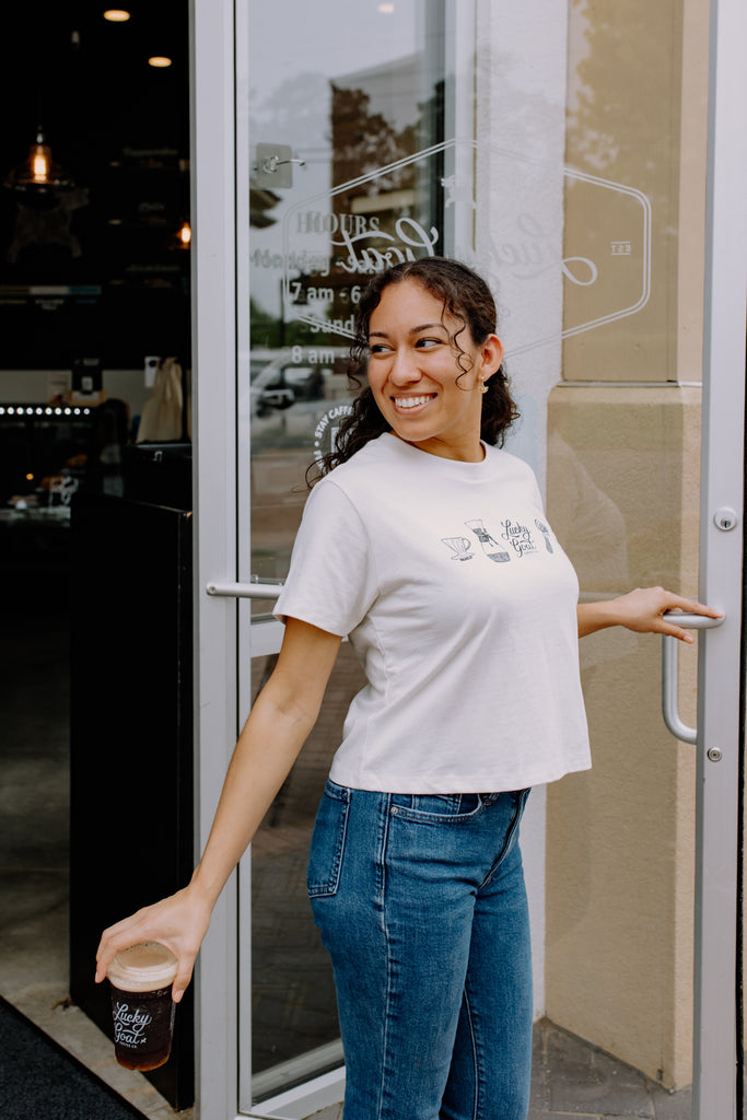 Woman in a white t-shirt and blue jeans holding a coffee cup, standing in front of a glass door.