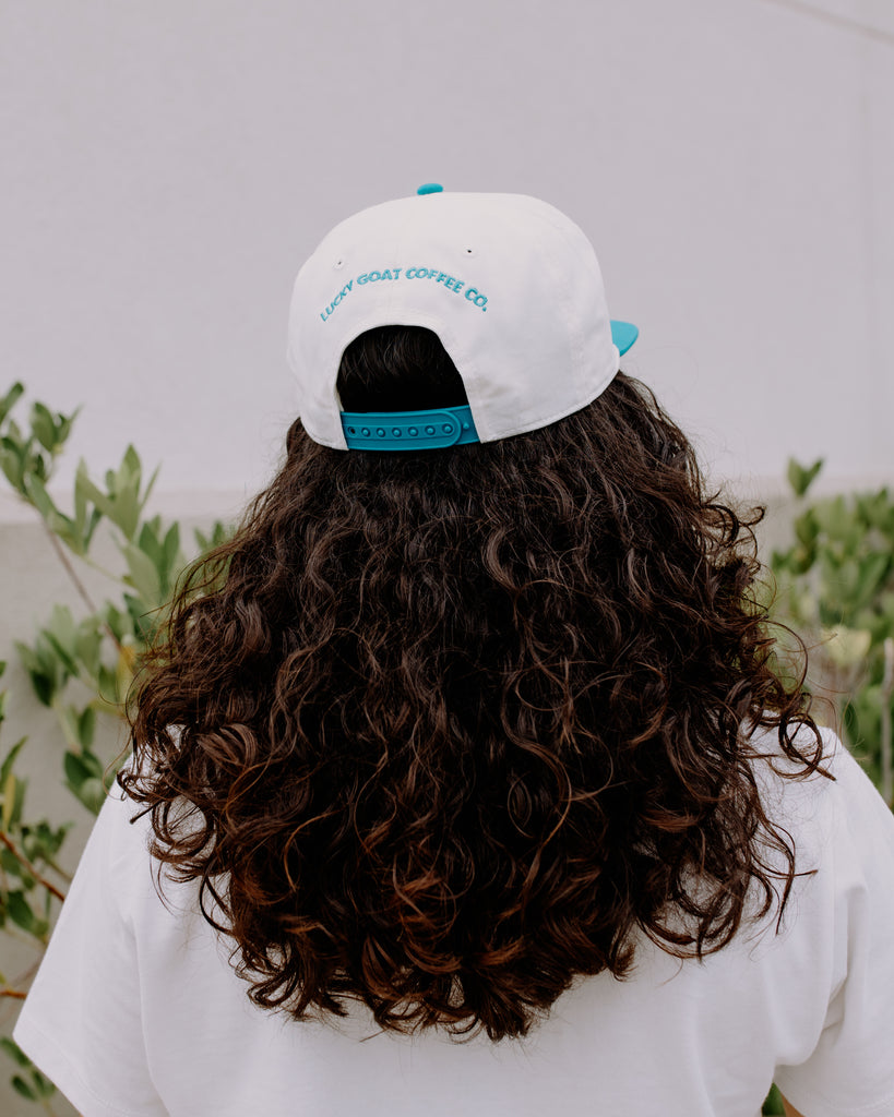 Person wearing a white cap with blue accents and text, standing outdoors with greenery in the background.