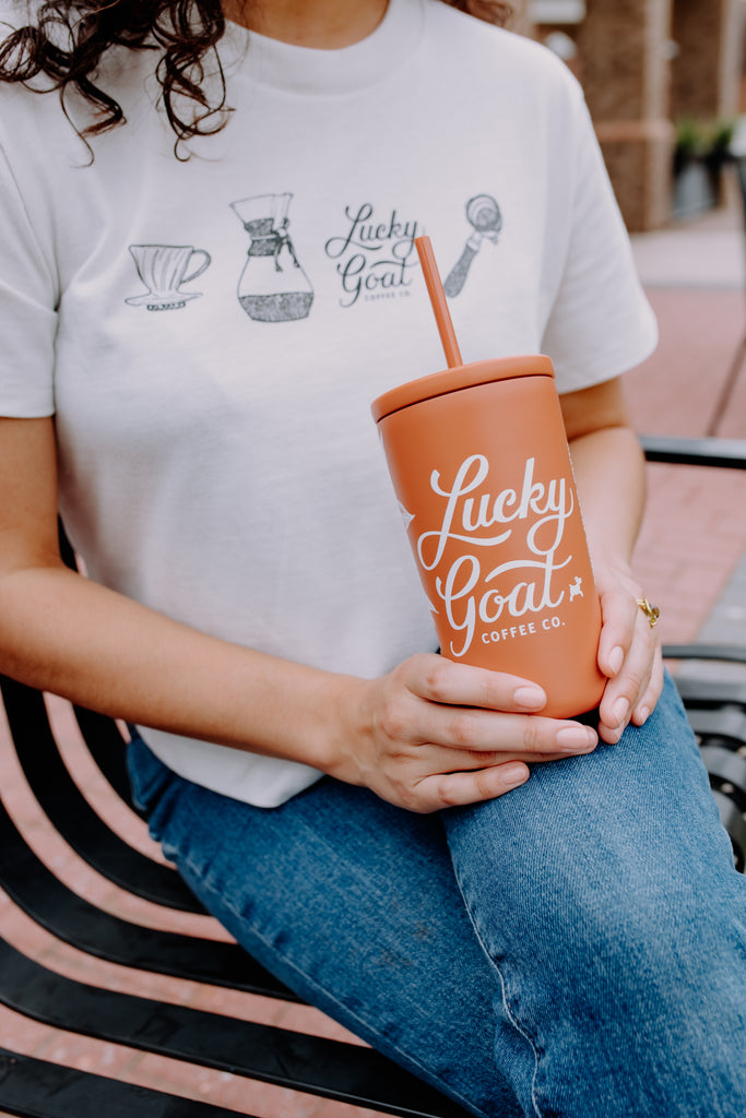 Person wearing a white t-shirt with coffee-themed designs and holding a 'Lucky Goat Coffee Co.' cup.