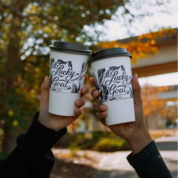 Two hands holding Lucky Goat hot coffee cups cheersing in the air with colorful foliage blurred in the background.