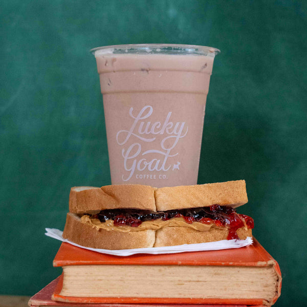 A cup of iced coffee with milk and a visible Lucky Goat Coffee Co. logo, styled on top of two orange books with a peanut butter and jelly sandwich in front, against a dark green chalkboard-like background.