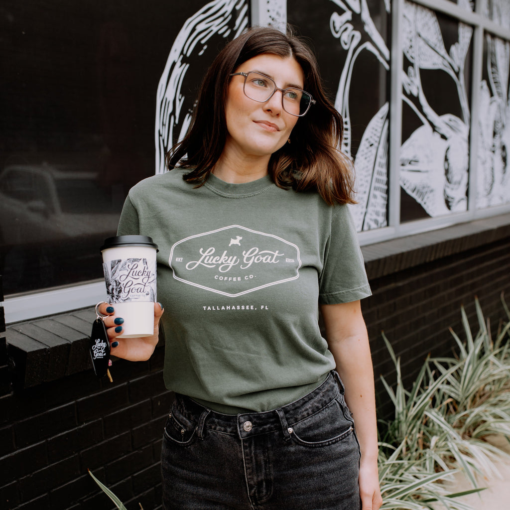 Person wearing a green t-shirt with a logo, holding a coffee cup, standing in front of a black and white mural.