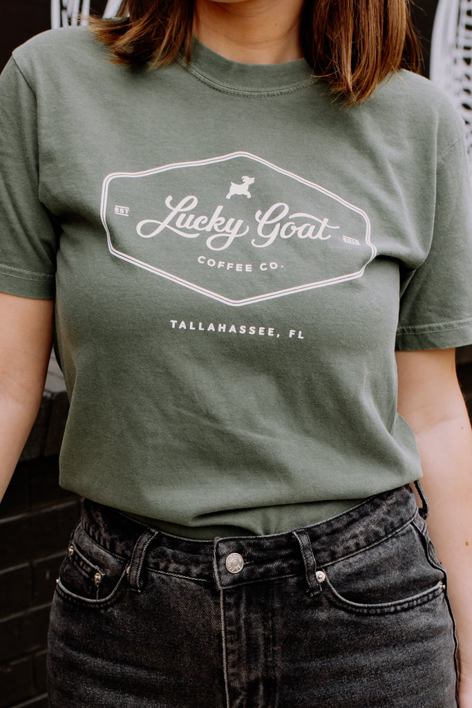 Person wearing a green t-shirt with a logo and holding a coffee cup, standing outdoors.
