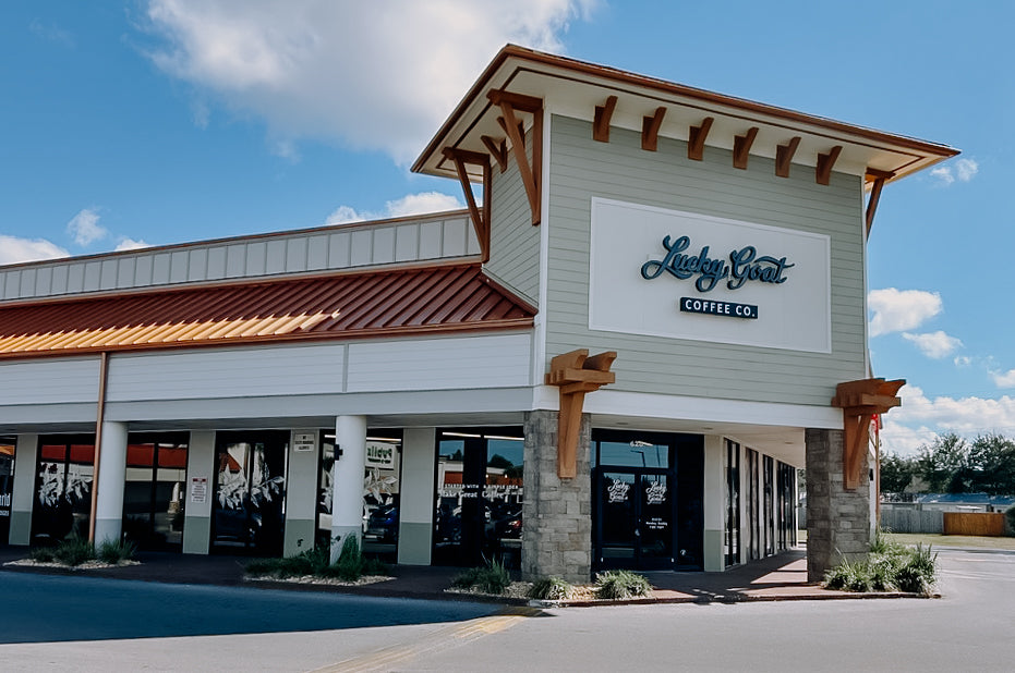 Lucky Goat Coffee Co. storefront under a blue sky, featuring light gray siding, a copper-colored metal roof, stone columns, and a prominent white sign with the brand name in teal script.