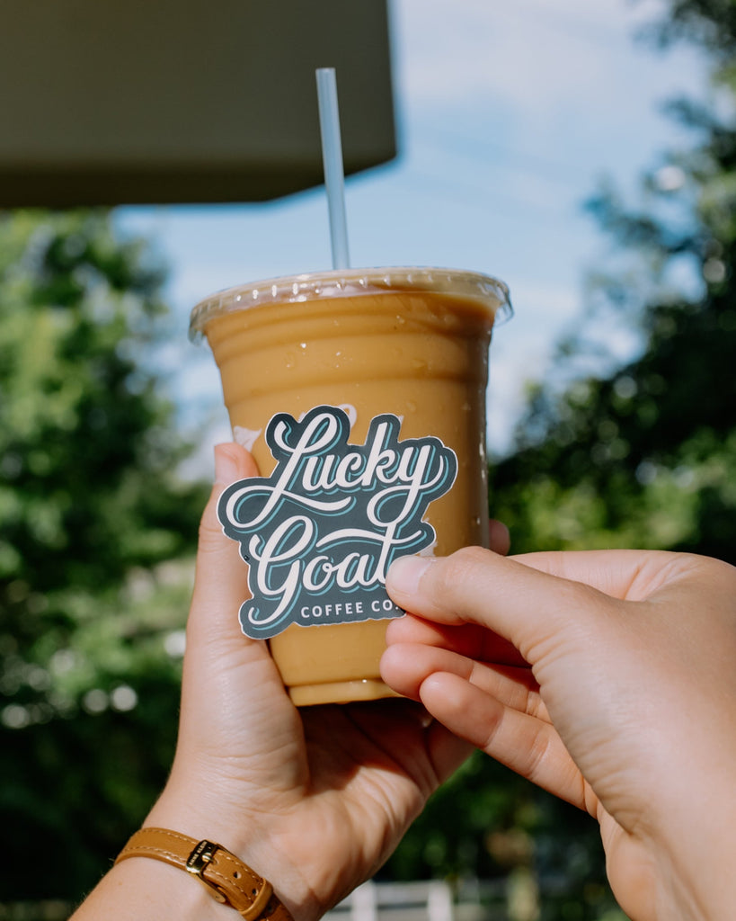 Person holding a cup of iced coffee with 'Lucky Goat Coffee Co.' label outdoors.
