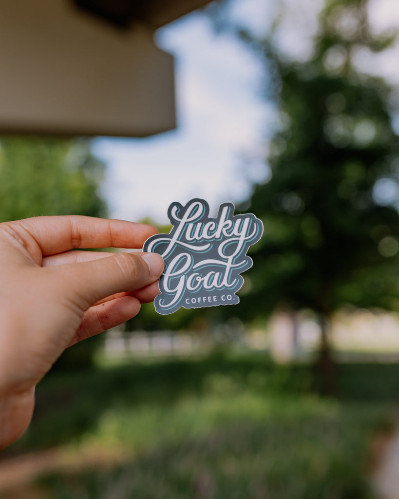 Hand holding a 'Lucky Goat Coffee Co.' sticker with a blurred outdoor background