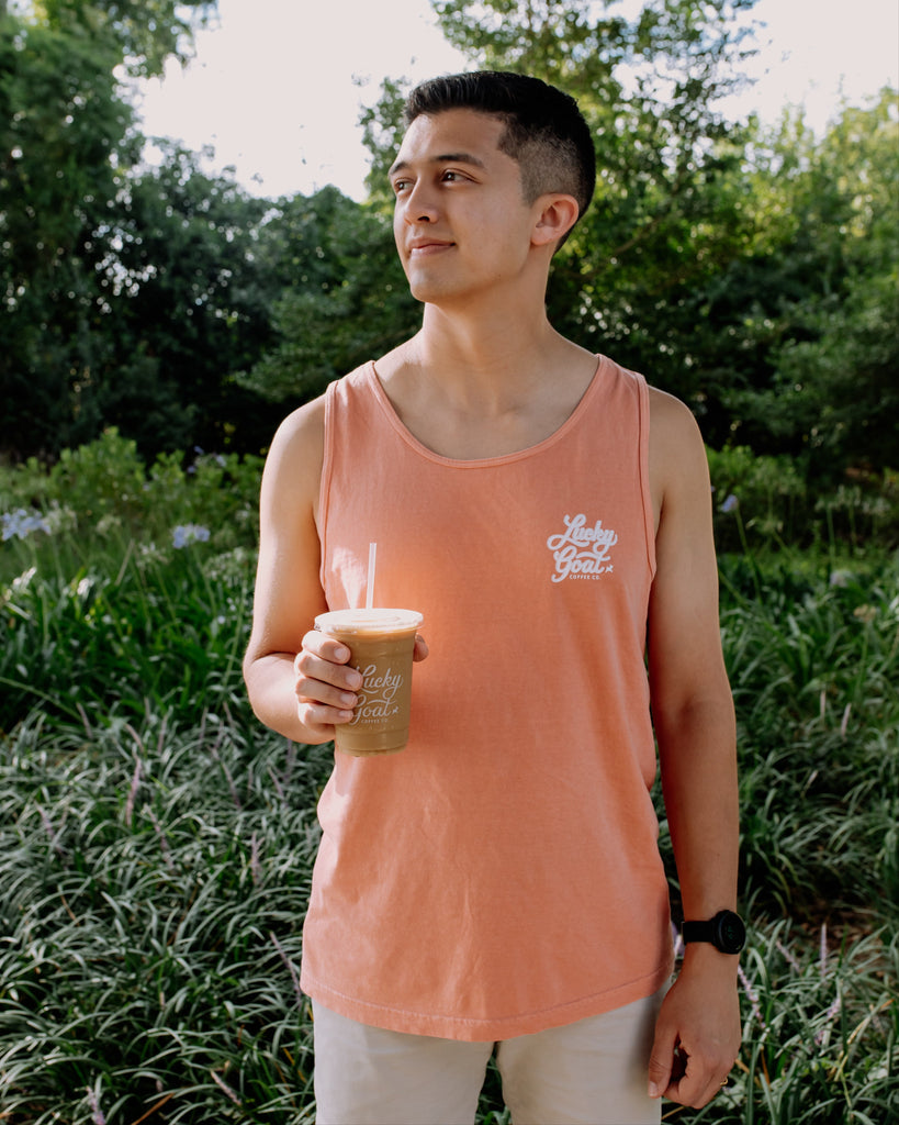 Man in a terracotta tank top holding a coffee cup outdoors with greenery in the background